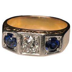 This is an Art Deco 14K Yellow Gold, Diamond and Sapphire Garter Ring. It depicts a gold ring with a top divided by two small squares at the sides and one larger one in the center. The center one is embellished by an old European brilliant diamond ( Weight- .75 carats; Measurements- 5.10 x 5.0 x 2.94 mm; Color- G; Clarity- SI-2; Type- Kimberly) and the side ones by two sapphires each one is weighing 0.5 carats. The ring is also decorated with an engraved branch of scrolls at the front and back o Square Cut Diamond Accent Rings In 14k Gold, 14k Gold Rings With Diamond Accents, Square Cut, 14k Gold Princess Cut Sapphire Ring With Brilliant Details, Square Cut 14k Gold Rings With Diamond Accents, 14k Gold Square Cut Single Diamond Ring, Art Deco Gold Three Stone Diamond Ring, Art Deco Three Stone Gold Diamond Ring, Gold Three Stone Art Deco Diamond Ring, Rectangular Three Stone Diamond Jewelry