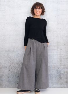 a woman standing in front of a white wall wearing wide legged pants and black top