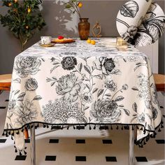 a table covered with a white and black flowered tablecloth next to a potted plant