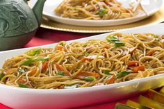 two white plates with noodles and vegetables in them on a red table cloth next to a green teapot