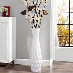 a white vase filled with lots of flowers on top of a wooden floor next to a window