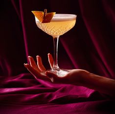 a woman's hand holding a cocktail glass with an orange peel on it, against a purple background