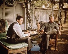 two men sitting on wicker chairs outside in the sun, one is eating food