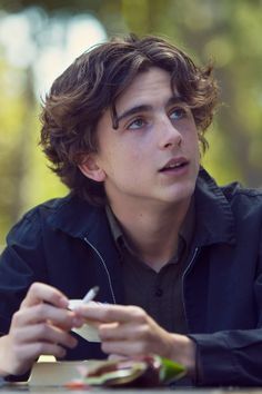 a young man sitting at a table with food in his hand and looking off into the distance