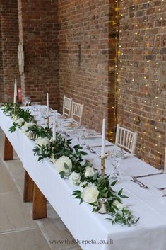 a long table with white flowers and greenery is set up in front of a brick wall