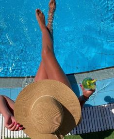 a woman in a straw hat is sitting on the edge of a swimming pool and holding a drink