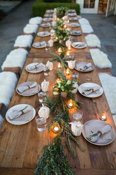 a long wooden table topped with white plates and greenery next to candles on top of it