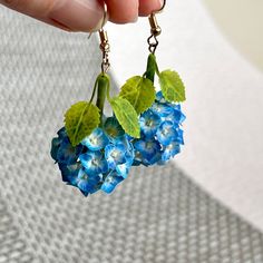 a person holding onto some blue flowers with green leafy stems hanging from it's ear wires