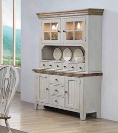 a white china cabinet with glass doors and two plates on the top, in front of a dining room table