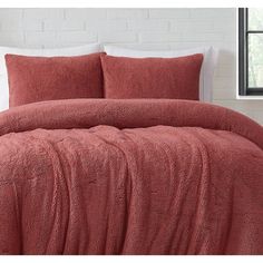 a bed with red sheets and pillows in front of a white brick wall that has a window