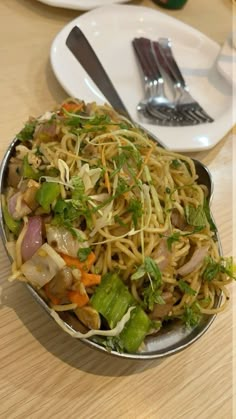 a bowl filled with noodles and veggies on top of a wooden table next to silverware