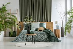 a bed with green sheets and pillows in a room filled with potted plants on the floor