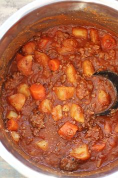 a pot filled with stew and potatoes on top of a wooden table next to a spoon