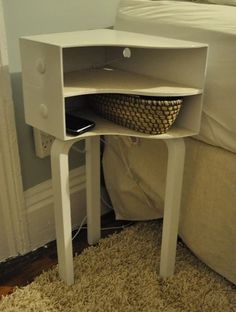 a small white table with a basket underneath it on the floor next to a bed