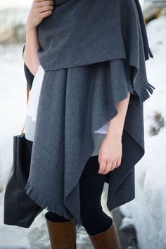 a woman is standing in the snow wearing a gray poncho and brown boots