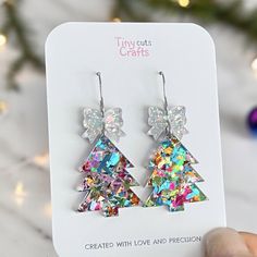 Christmas Tree Glitter Earrings adorned with a sparkling Holographic bow. Expertly crafted from glitter in vibrant shades of pink, silver, green, and blue, these earrings feature an eye-catching acrylic bow for an extra dash of holiday cheer. Measuring 1.75 to 2 inches in height and 1.20 inches in width, they are the perfect accessory to celebrate the season. Ideal as a Christmas gift for her, these earrings bring festive elegance and charm to any ensemble. Arriving in a gift bag accompanied by White Jewelry For Holiday Party, White Holiday Party Jewelry, Silver Holiday Party Jewelry, Silver Jewelry For Holiday Parties, Silver Jewelry For Party And Holiday Season, Silver Jewelry For Party And Holiday, Holiday Multicolor Earrings As A Gift, Holiday Multicolor Earrings Perfect For Gifts, Holiday Multicolor Earrings As Gift