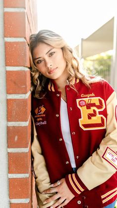 a woman leaning against a brick wall wearing a varsity jacket