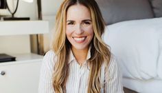 a smiling woman sitting in front of a bed