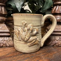 a ceramic mug with a frog design on the side sitting on a wooden table next to a potted plant