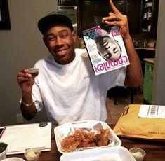 a man holding up a magazine over his face while sitting at a table full of food