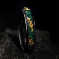 a green and gold wedding band with an intricate design on it's side, sitting on top of a rock