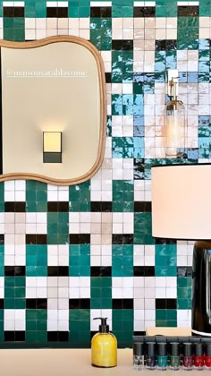 a bathroom with green and white tiles on the wall next to a lamp, mirror and soap dispenser