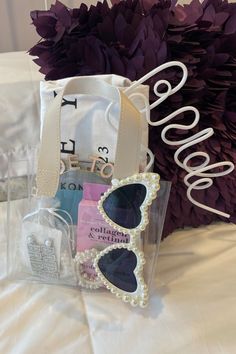 a bag with sunglasses and some other items in it on a bed next to purple flowers