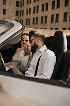 a man and woman sitting in the back seat of a car, one holding up her sunglasses