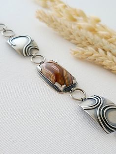 Reminiscent of steep desert canyon walls, slowly cut away by meandering waterways, this bracelet is purely inspired by the layered bands of color winding through the Laguna Lace Agate. All the hues of the desert can be found in this unique stone: red clay, faded green, sandy browns, and sunset purples. The stone is held in place with silver at all four corners so you can see the colors moving through the agate, and a window cut in the back reveals the continuous maze of colorful patterns on the Artisan Cabochon Bracelet Jewelry, Bohemian Sterling Silver Oyster Bracelet, Artisan Agate Jewelry With Polished Finish, Handmade Jasper Bracelet, Brown Polished Agate Jewelry, Modern Natural Stones Bracelet, Unique Agate Gemstone Bracelets, Bohemian Jasper Bracelet, Modern Natural Stone Bracelet