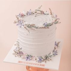 a white cake with purple flowers and greenery on top is sitting on a stand