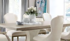 a dining room table with white chairs and a vase on top of it in front of a window