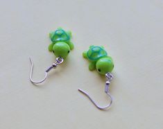 two green turtle shaped earrings with hooks on a white surface, one is dangling from the hook