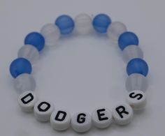 Show off your team pride with this handmade Dodgers beaded bracelet, a must-have accessory for any true fan! This bracelet features alternating frosted blue and white beads, perfectly representing the iconic colors of your favorite baseball team. The centerpiece of this bracelet is the bold "DODGERS" lettering, crafted with high-quality white beads and clear black lettering, ensuring your allegiance is always front and center. Whether you're cheering from the stands, hosting a game day party, or simply showing your support around town, this bracelet is the perfect way to display your love for the Dodgers. It's made with durable elastic cord, allowing it to stretch comfortably to fit most wrist sizes, so you can wear it all season long without missing a beat. This bracelet makes an excellen White Team Spirit Stretch Bracelet, Personalized Blue Bracelets For Game Day, White Team Spirit Stretch Bracelet As Gift, Blue Adjustable Bracelets For Team Spirit, Blue Team Spirit Beaded Bracelets As Gift, Personalized White Wristband Team Spirit, Blue Beaded Bracelets With Letter Beads For Team Spirit, Blue Beaded Bracelets For Team Spirit Gift, Blue Adjustable Beaded Bracelets For Team Spirit