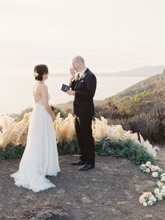 a bride and groom standing on top of a hill looking at each other with flowers in front of them