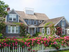 a white picket fence with pink flowers in front of a large gray house and trees