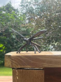 a small metal insect sitting on top of a wooden bench in front of some trees