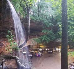 a waterfall in the middle of a forest with people sitting at tables under it and trees around