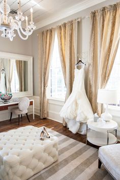 a wedding dress hanging on a hanger in front of a window with curtains and chandelier