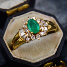 an emerald and diamond ring sitting on top of a velvet covered box with gold trimmings