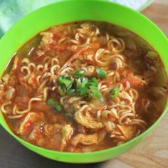 a green bowl filled with noodles and meats on top of a wooden cutting board