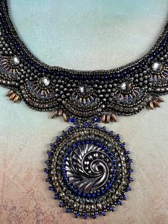 an elaborate necklace with blue beads and silver accents on a white surface, in the shape of a peacock