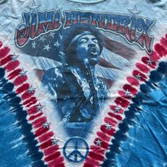 Step into the music scene with this Vintage Jimi Hendrix American Flag Blues Guitarist Tie Dye Tee. Officially licensed and medium-sized, this shirt is a genuine tribute to the legendary guitarist. Its tie-dye pattern with the American flag motif channels the hippie vibes of the era. Ideal for music enthusiasts, collectors, and those who appreciate vintage fashion, this tee is more than apparel; it's a piece of concert memorabilia representing a crucial moment in music history. Whether you're a Blue Screen Print T-shirt For Concert, Blue Band Merch T-shirt, Band Logo Cotton Tops For Festival, Cotton Band Logo Tops For Festival, Rock And Roll Screen Print Tops For Music Festivals, Cotton Tops With Band Logo For Festival, Band Logo Cotton T-shirt For Festival, Cotton T-shirt With Band Logo For Festival, Pop Culture Crew Neck Top For Music Festivals