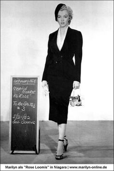 a woman in a black suit and heels standing next to a chalkboard with writing on it