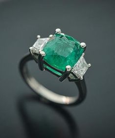 This Elegant Platinum three stone ring has a Natural Cushion Cut Colombian Emerald from the Top Muzo Mine with a GIA Certificate. The side stones are Natural Brilliant Cut Trapezoid Pair of Matching Diamonds. GIA#5202111099 3.34 Carat Cushion Modified 9.64x8.93x5.64 with moderate F2 clarity enchancement. 2 Matching Trapezoid Brilliant Cut Diamonds weigh 0.71 carats total weight and are graded G in color and SI1 to SI2 in clarity. Platinum weight is 5.14 DWT. Ring can be sized. Luxury Emerald Three Stone Ring For Formal Occasions, Luxury Three Stone Emerald Ring For Formal Occasions, Luxury Three Stone Emerald Ring For Formal Events, Luxury Three Stone Emerald Ring, Luxury Three Stone Gemstones For Anniversary, Luxury Three Stone Asscher Cut Emerald Ring, Luxury Three Stone Emerald Jewelry, Luxury Three-stone Emerald Ring, Luxury Asscher Cut Three Stone Emerald Ring