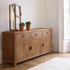 a wooden cabinet with two potted plants on top and a mirror above it in a white room