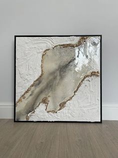 a white and brown abstract painting in a black frame on the floor next to a wall