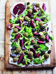 a wooden cutting board topped with salad and berries next to a bowl of dressing on top of it