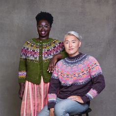 two women sitting next to each other in front of a gray wall wearing colorful sweaters