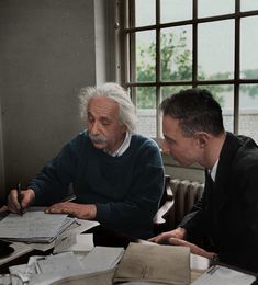 two men sitting at a table with papers and pens in front of them, one is writing