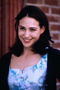 a smiling woman in a blue dress and black cardigan standing next to a brick wall